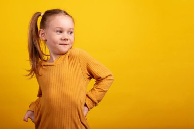 Hardallı süveter giyen genç bir kız, elleri kalçasında, şakacı bir şekilde omzunun üstünden bakıyor, parlak sarı bir arka planda..