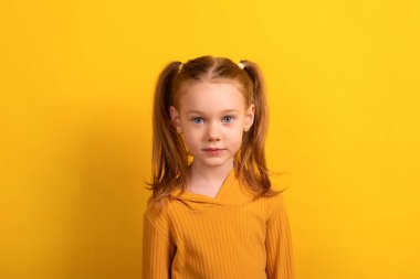 Saçları örgülü, hardal sarısı süveter giyen genç bir kız. Saf sarı bir arkaplanda duruyor ve doğrudan kameraya bakıyor..