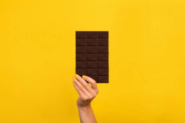 Sarı ojeli kadın eli parlak sarı arka planda koyu çikolatayı tutuyor. Kopyalama alanı olan minimalist kompozisyon.