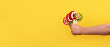 Çocuğun eli, parlak sarı bir zemin üzerinde çubuklar üzerinde canlı meyve şekilli lolipoplar tutuyor, neşe ve tatlılığı çağrıştırıyor..