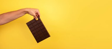 Sarı ojeli bir kadın büyük siyah bir çikolatayı parlak sarı bir arka plana dikey olarak tutuyor..