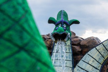 Kaprisli yeşil bir ejderha çeşmesinin yakın çekimi. Ağzından su akıyor, açık havada bulutlu bir gökyüzüne karşı..