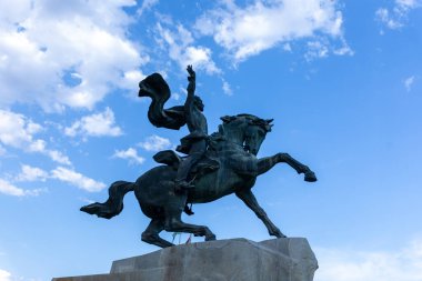 Dinamik binicilik heykeli, parlak mavi gökyüzüne karşı dağınık bulutlarla, gücü ve liderliği sembolize ediyor. Tarih, kültür veya ilham temaları için ideal.