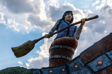 Süpürge tutan renkli bir cadının heykeli, bir fıçının üzerine oturmuş, bulutlu bir gökyüzüne kurulmuş. Folklor, Cadılar Bayramı veya fantezi teması için mükemmel..