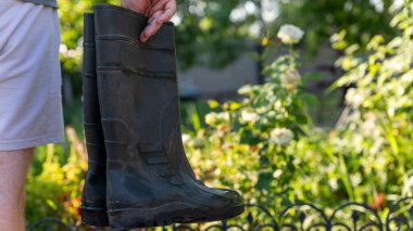 Siyah lastik çizmeleri olan, arka planında bulanık yeşil bir bahçe olan ve çiçek açan birinin yakın çekimi..