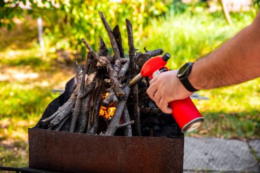 Barbekü ızgarasında odun tutuşturan bir adam açık havada kırmızı bir fener çakmağı ile, yeşil doğa ve yaz güneşiyle çevrili..