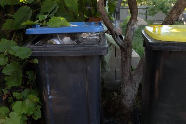 İki büyük açık hava çöp kutusu, birinin kapağı atıkla dolu, yerleşim alanındaki yeşilliklerle çevrili..