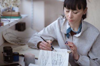Kadın ressam çizimlerine kalem ve siyah mürekkeple bakıyor. Grafik boyama. Modern sanat yaratılıyor
