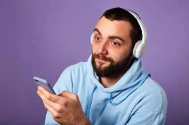 Ciddi sakallı adam beyaz kulaklıkla müzik dinliyor ya da podcast yapıyor. Telefon kullanılıyor