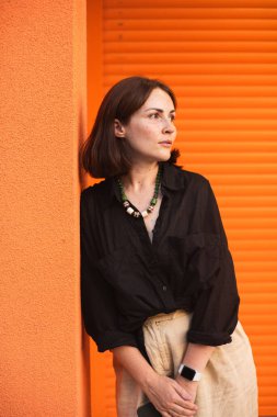 Sıradan kıyafetlerle turuncu duvara yaslanan bir kadın. Kameraya gülümsüyor..