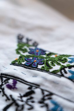 Geleneksel bir zanaat ortamında beyaz kumaş zemin üzerinde mor çiçekler ve yeşil asmalar bulunan el yapımı nakış. Geleneksel Ukrayna Nakışı