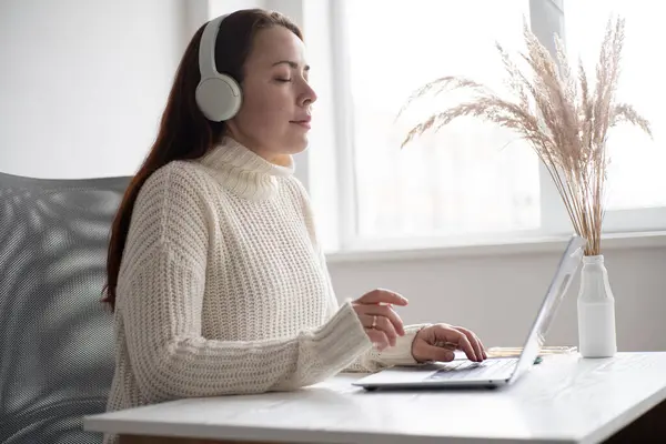 Aydınlık bir odada bitkilerle otururken kulaklık takan bir kadın dizüstü bilgisayarla çalışıyor.