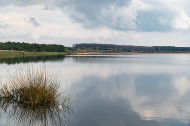Sakin göl bulutlu gökyüzünü yansıtır ve kıyıda yürüyen insan siluetleri doğal manzara içinde.