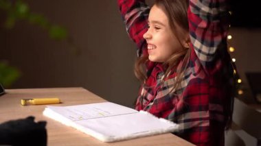 Küçük bir çocuk ödevlerini rahat bir masada, iyi aydınlatılmış bir odada tamamlayıp heyecan ve başarı göstererek kutluyor.