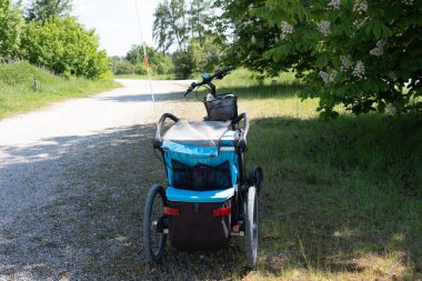 Mavi bir bisiklet römorku, çevresi yeşilliklerle çevrili, sakin bir çakıl yolunda durur. Açık hava seyahatinin ve maceranın özünü yakalar..