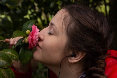 Kırmızı ceketli bir kadın pembe kamelya çiçeğini tutar ve koklar, gözleri kapalı, doğanın barışçıl ve duyusal güzelliğini kucaklar..