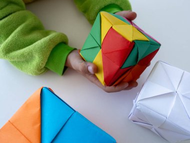 Yeşil kollu çocuk beyaz masada parlak modüler origami küpü tutuyor..