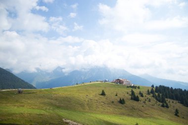 Parlak bulutlu bir gökyüzünün altında, orman ve tepelerle çevrili yeşil bir dağ evi..