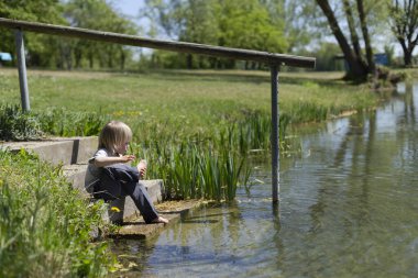 Çıplak ayaklı çocuk taş basamaklarda oturuyor, güneşli bir günde ayaklarını gölete daldırıyor..