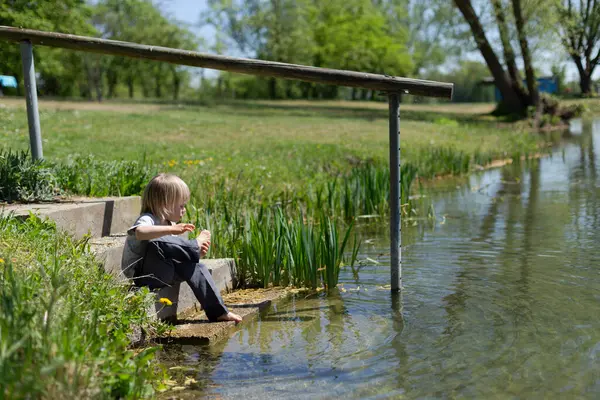 Çıplak ayaklı çocuk taş basamaklarda oturuyor, güneşli bir günde ayaklarını gölete daldırıyor..