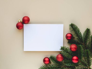 Tarafsız bir bej arka planda boş bir beyaz kartı çerçeveleyen kırmızı Noel süsleri ve çam dallarının yer aldığı bir şenlik düzenlemesi. Bayram tebrikleri, Noel kartları veya mevsimlik promosyonlar için ideal..