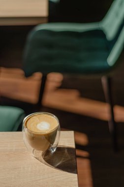 Sıcak, davetkar bir fincan kahve, rahat bir kafe ortamında, dolgun yeşil sandalyelerle çevrili ahşap bir masanın üzerinde oturur. Yumuşak ışıklandırma ve minimalist dekor rahatlatıcı bir ortam yaratır, mükemmel.