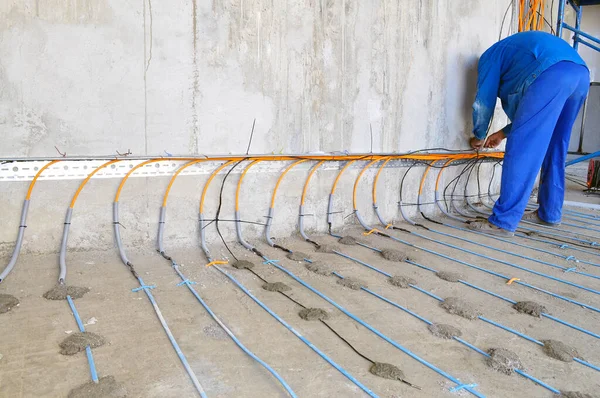A worker in a blue uniform is engaged in laying a floor heating cable on concrete, a man from the back