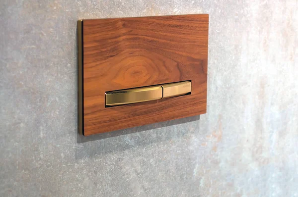Button for wall-hung toilet installation, wood texture imitation with gold accents on a backdrop of gray concrete tiles. Vibrant architectural design