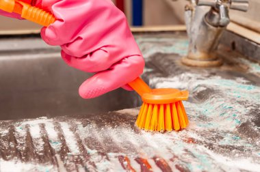 Kirli ve sert su lekelerini temizlemek için kirli bir mutfak lavabosunu toz temizleyici ve fırçayla ovalayan eldivenli bir elin yakın çekimi..