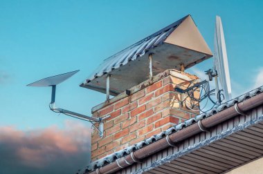 Düşük açılı çekimde metal kapaklı tuğla baca, uydu anteni ve bir evin tepesine yerleştirilmiş Starlink internet alıcısı görülüyor..