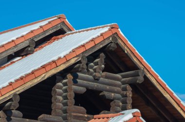 Kırsal bir ahşap kulübeye yakın çekim, terracotta kiremit çatılı, parlak bir kış gününde açık mavi gökyüzüne karla kaplanmış.