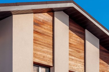 Parlak mavi gökyüzüne karşı hafif ahşap kaplamalı ve tarafsız sıva kolonları olan modern bir evin dış görünüşüne yakın çekim. Mimari ayrıntı.