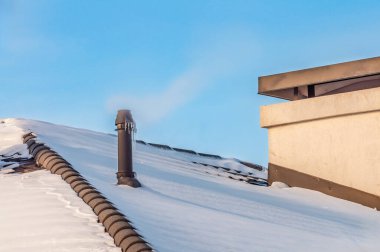 Kışın, karla kaplı bir çatıda kışın serinliğini ve mimari detaylarını gözler önüne seren açık, mavi bir gökyüzüne buhar yayan çatı havalandırması manzarası..
