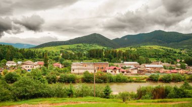 Bereketli yeşil tepeler ve bulutlu bir gökyüzünün altında akan bir nehir arasında yuva yapmış tuhaf bir köy manzarası, seyahat ve doğa temaları için mükemmel..