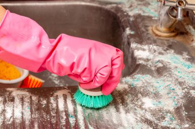 Pembe eldiven giyen biri eski bir lavaboyu temizlik malzemesi ve yeşil bir fırçayla temizliyor..