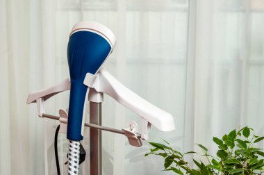 Üzerinde elbise askısı olan mavi ve beyaz bir buharlı buharlı buharlı tencerenin yakın çekimi. Perdeli, yeşil yapraklı bitkinin yanında..