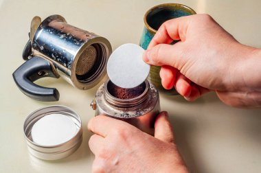 Moka demliği kullanarak kahve yapan biri. Taze bir fincan demlemeden önce kağıt filtre koyuyorlar, arka planda kupa var..