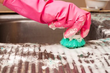 Eldivenli bir el, lekeleri temizlemek için yeşil bir fırça ve sabunlu suyla ovalıyor. Temizlik ya da ev işlerini işaret ediyor..