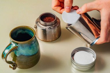 Daha yumuşak, daha az acı bir bira için kağıt filtreli moka otu kahvesi hazırlıyorum. Tarafsız arkaplanda Rustik seramik bardak ve alüminyum kap.