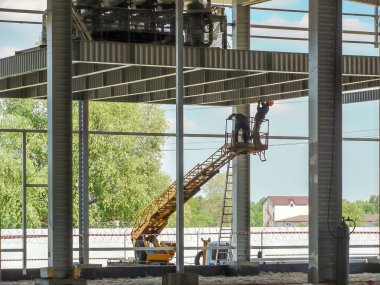 Sarı bir makas asansöründeki iki inşaat işçisi yeni bir sanayi binası çerçevesinde çelik çatı kirişleri kuruyor..