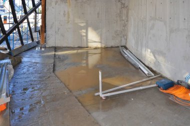 İnşaat sahasının beton zemininde duran su, gün ışığını yansıtır. Cihazlar ve malzemeler, etkin çalışmaların devam ettiğini gösterir..