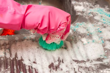 Pembe eldiven giyerken yeşil bir fırçayla sabun ve deterjanla kaplanmış bir yüzeyi ovalayan birinin yakın çekimi..