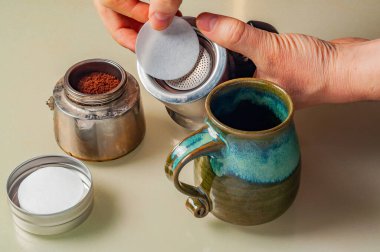 Görüntüde Moka demliği ile kahve hazırlayan ve filtre kağıdı yerleştiren bir kişi yer alıyor. Seramik bir bardak, sıcak kahve demleme işini tamamlar..