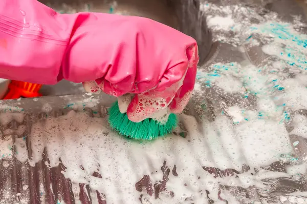 Pembe eldiven giyerken yeşil bir fırçayla sabun ve deterjanla kaplanmış bir yüzeyi ovalayan birinin yakın çekimi..