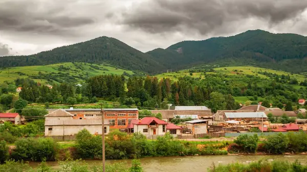 Ukrayna Karpatlarının yemyeşil tepeleri, geleneksel binaları ve bulutlu bir gökyüzünün altında akan bir nehri olan manzarası..