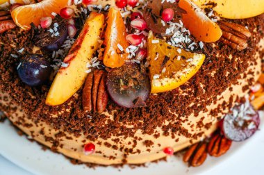 Portakal, üzüm ve şeftali de dahil olmak üzere çeşitli taze meyvelerle süslenmiş güzel bir pasta. Pasta fındık ve hindistan cevizi parçacıklarıyla kaplı..