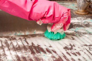 Pembe koruyucu eldiven giyen bir insan, yakın plan çekimde kirli bir metal yüzeyi sabunlu su ve kirle kaplamak için yeşil bir fırça kullanır..