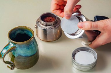 Yakın plan çekimde kahve makinesi, filtre kağıdı ve ev yapımı kahve kupası hazırlayan bir kişi görülüyor..