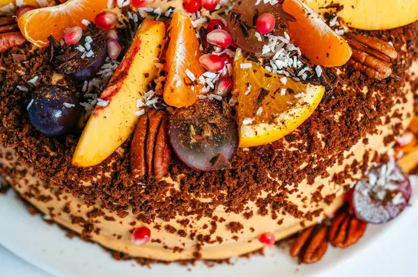 Portakal, üzüm ve şeftali de dahil olmak üzere çeşitli taze meyvelerle süslenmiş güzel bir pasta. Pasta fındık ve hindistan cevizi parçacıklarıyla kaplı..