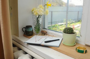 Sakin bir sahne, içinde bir vazo çiçek, bir defter ve bir kaktüs olan pencere pervazını gösteriyor. Olmak için güzel bir yer..